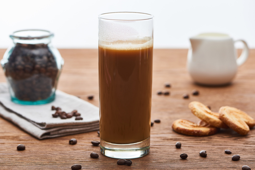 selective focus of ice coffee with milk in glass near milk jug, cookies and coffee grains on wooden table isolated on white - Photo, Image