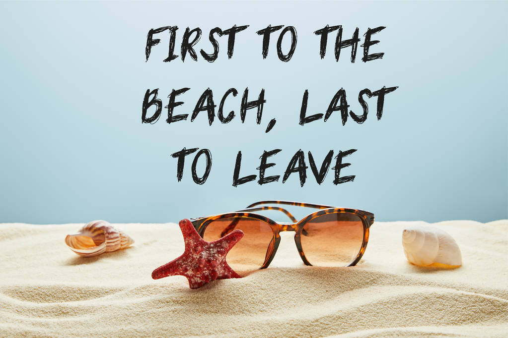 brown stylish sunglasses on sand with seashells and starfish on blue background with first to the beach, last to leave lettering - Photo, Image