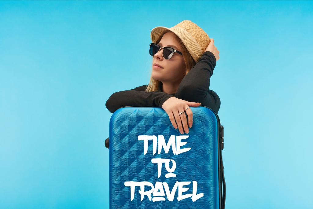 blonde girl in sunglasses and straw hat near blue suitcase with time to travel illustration isolated on blue - Photo, Image