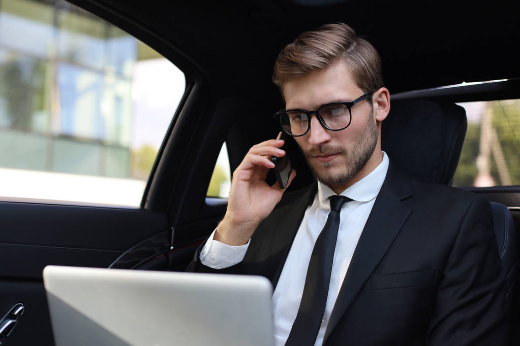 Knappe zelfverzekerde zakenman in pak praten op slimme telefoon en werken met behulp van laptop terwijl zittend in de auto. - Foto, afbeelding