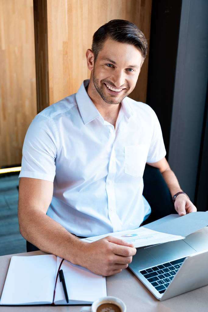 uśmiechnięty mężczyzna trzyma folder siedząc przy stole z laptopem w kawiarni - Zdjęcie, obraz
