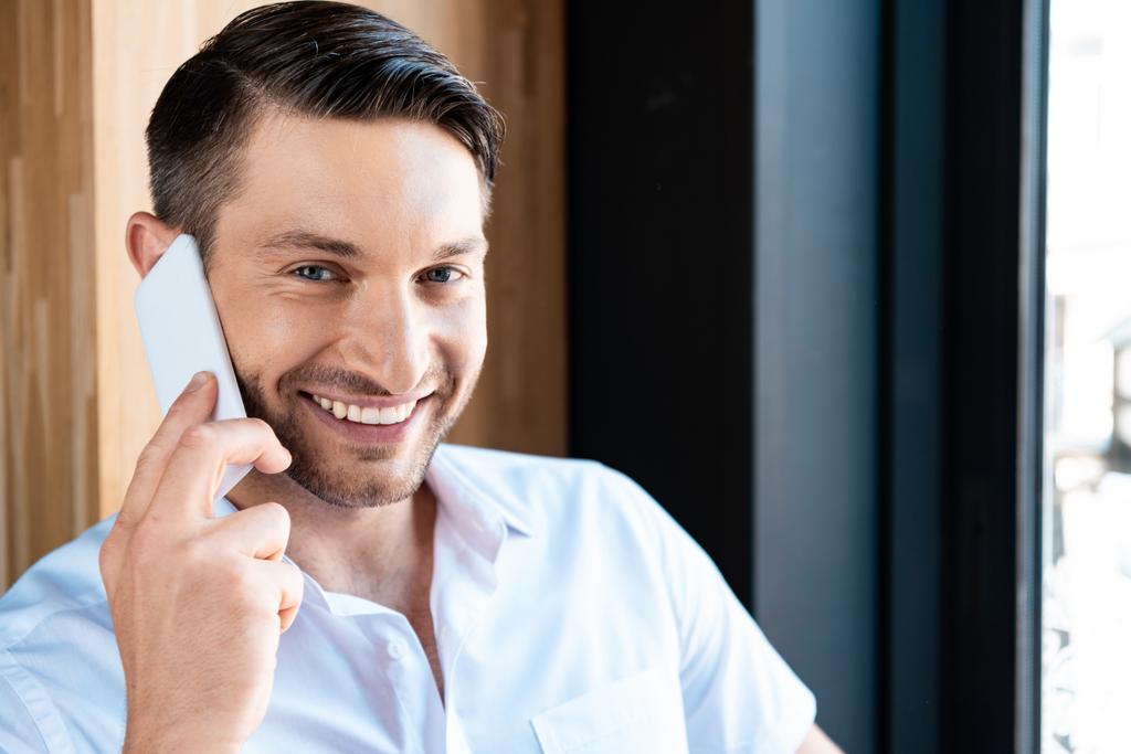 beyaz gömlekli gülümseyen adam akıllı telefonda konuşuyor ve kameraya bakıyor - Fotoğraf, Görsel