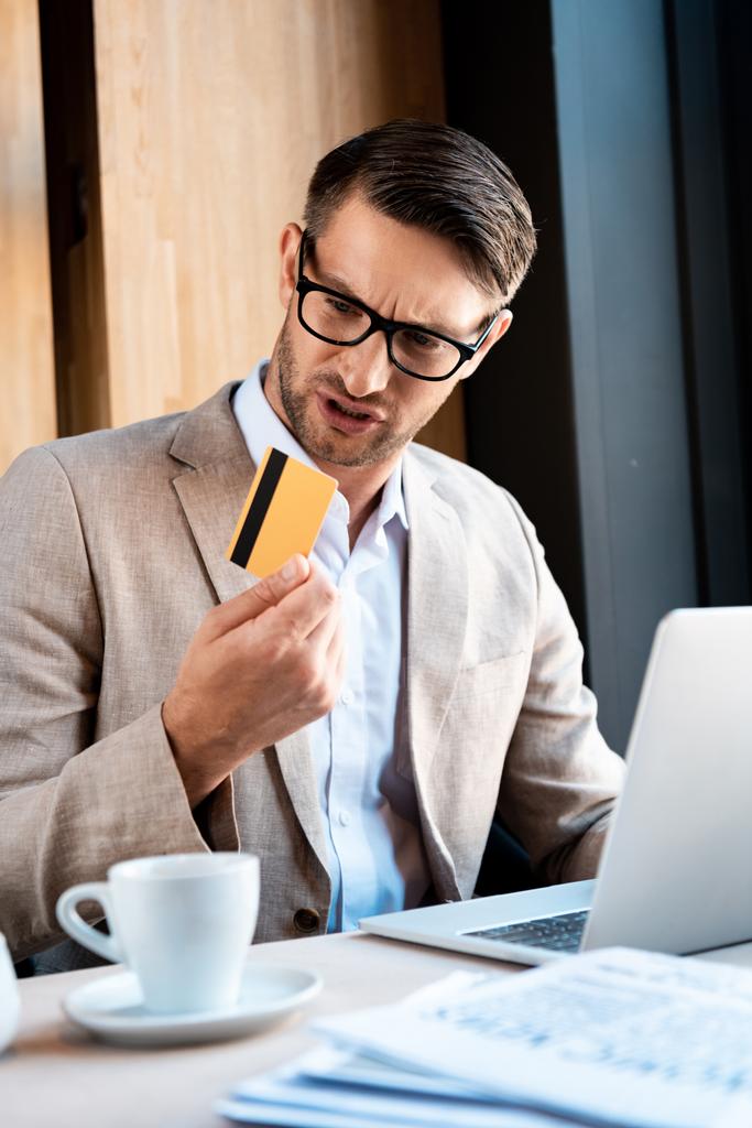 μπερδεμένος επιχειρηματίας σε ποτήρια με πιστωτική κάρτα και φορητό υπολογιστή στο καφέ - Φωτογραφία, εικόνα
