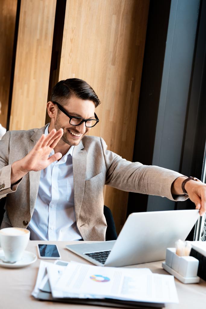 hombre de negocios emocionado en gafas agitando la mano a la pantalla del ordenador portátil en la cafetería
 - Foto, Imagen