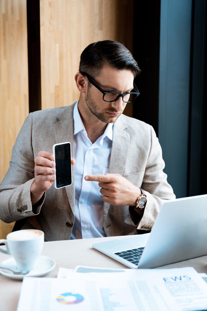 hombre de negocios señalando con el dedo en el teléfono inteligente con pantalla en blanco delante de la computadora portátil en la cafetería
 - Foto, imagen