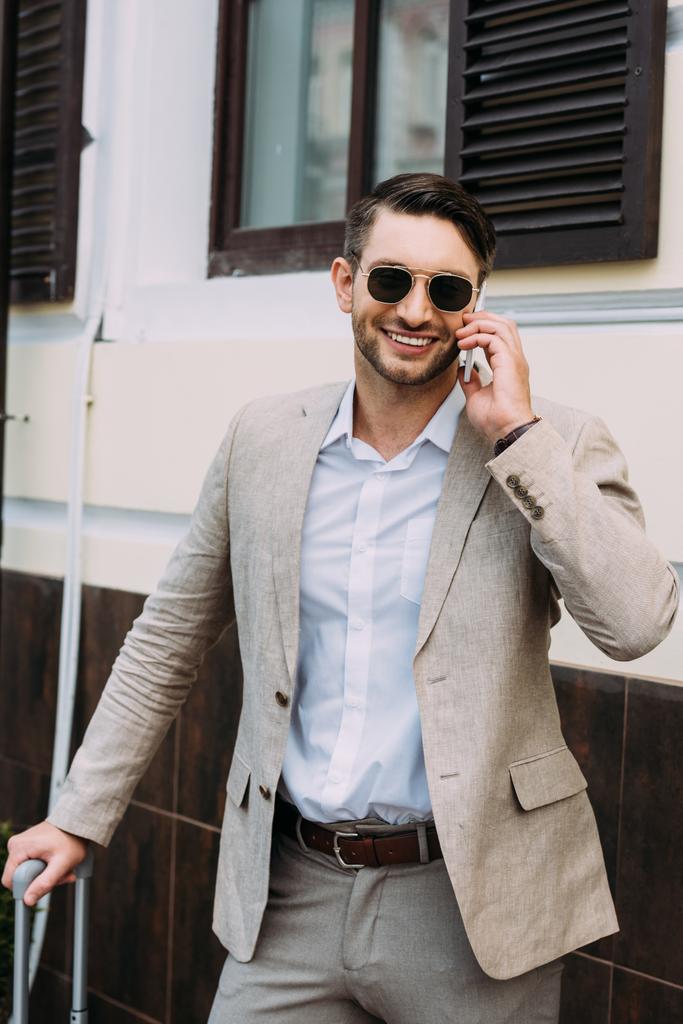 улыбающийся бизнесмен в солнечных очках, держащий чемодан и разговаривающий по смартфону на улице
 - Фото, изображение