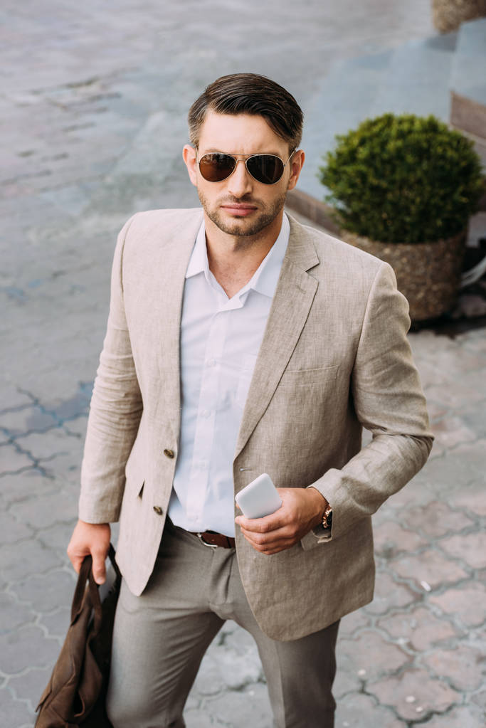 serieuze zakenman in zonnebril houden tas en smartphone op straat - Foto, afbeelding