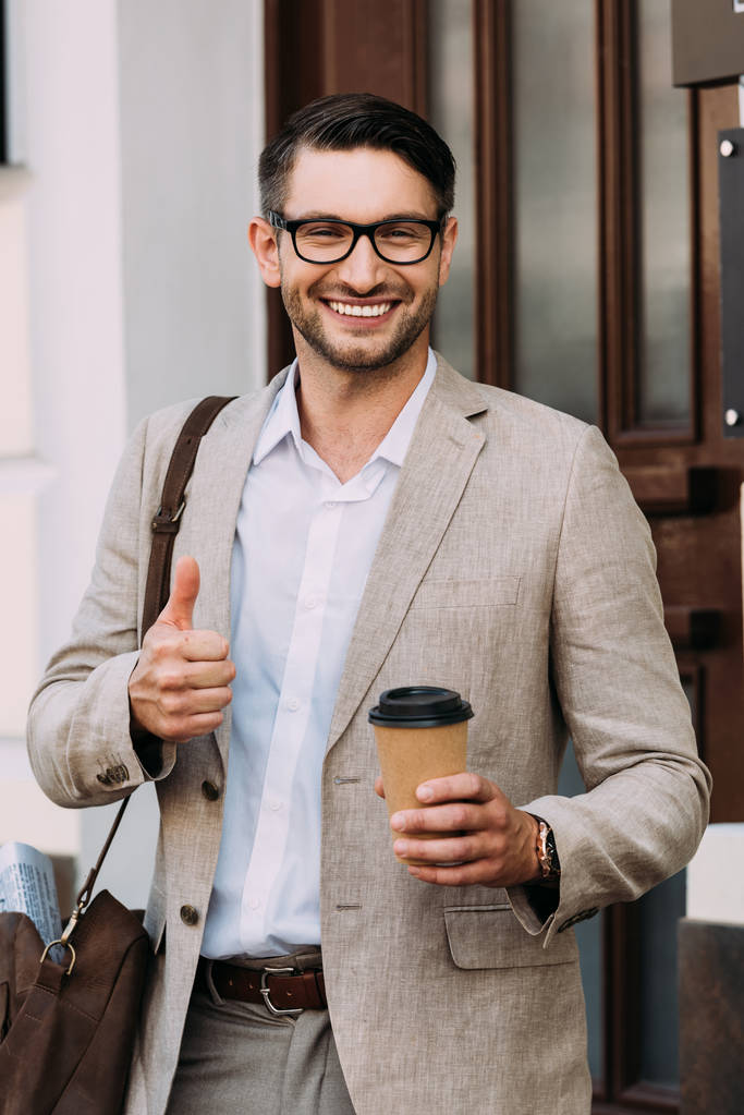 Vorderansicht eines lächelnden Geschäftsmannes mit einer Brille, die einen Coffee to go hält und den Daumen auf der Straße zeigt - Foto, Bild