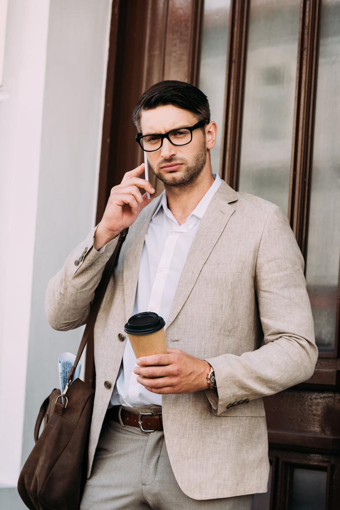 Nachdenklicher Geschäftsmann mit Ledertasche, der auf dem Smartphone spricht und Kaffee hält, um auf die Straße zu gehen - Foto, Bild