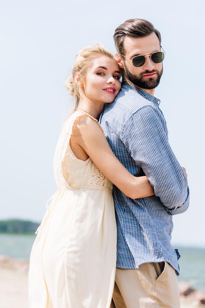 lage hoek uitzicht van gelukkige blonde vrouw knuffelen bebaarde vriendje op het strand - Foto, afbeelding