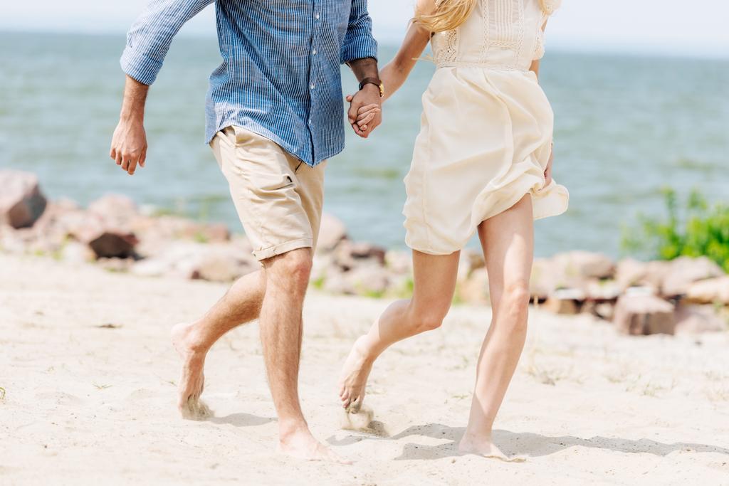 ビーチに沿って走りながら手をつないでいる裸足の若いカップルのトリミングされた眺め - 写真・画像