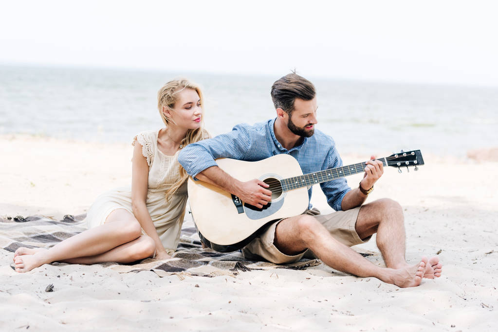 güzel genç yalınayak kadın denize yakın plajda akustik gitar çalan erkek arkadaşı ile battaniye üzerinde oturan - Fotoğraf, Görsel