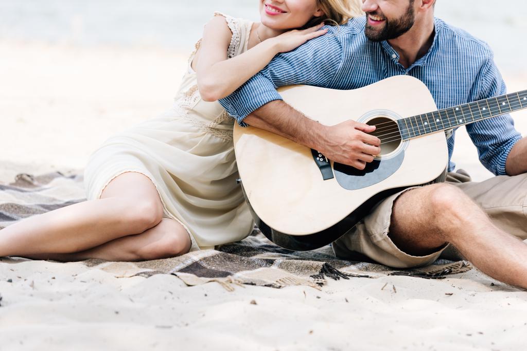 denize yakın plajda akustik gitar ile battaniye üzerinde oturan güzel genç çift kırpılmış görünümü - Fotoğraf, Görsel