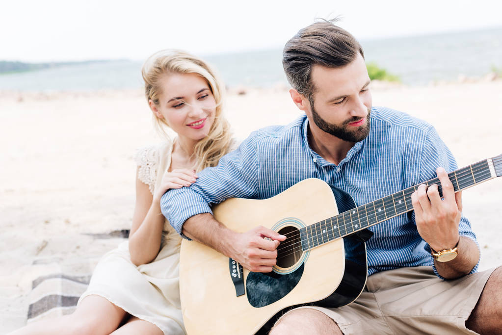fiatal szőke mezítláb nő ül takaró közeli barátja, akusztikus gitár a strandon a tenger közelében - Fotó, kép