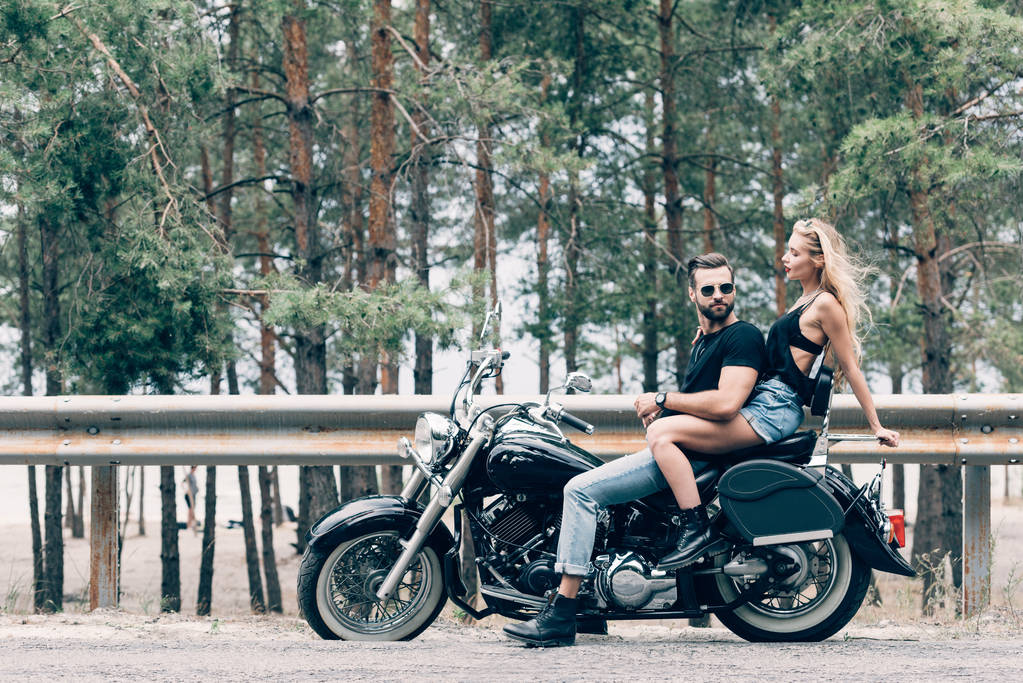 side view of young sexy couple of bikers on black motorcycle on road near green forest - Photo, Image