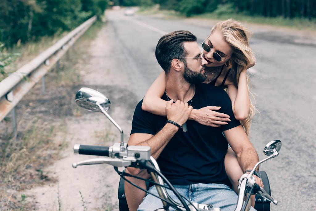 緑の森の近くの道路上の黒いオートバイでお互いを抱きしめ、お互いを見ているバイカーの若いカップル - 写真・画像