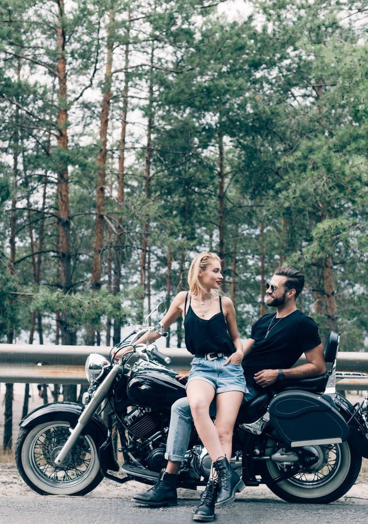 souriant jeune couple de motards assis et se regardant sur la moto noire sur la route près de la forêt verte
 - Photo, image