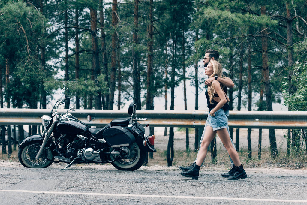 道路に沿って歩き、黒いオートバイと緑の森の近くに抱き合うバイカーの若いカップルの側面図 - 写真・画像