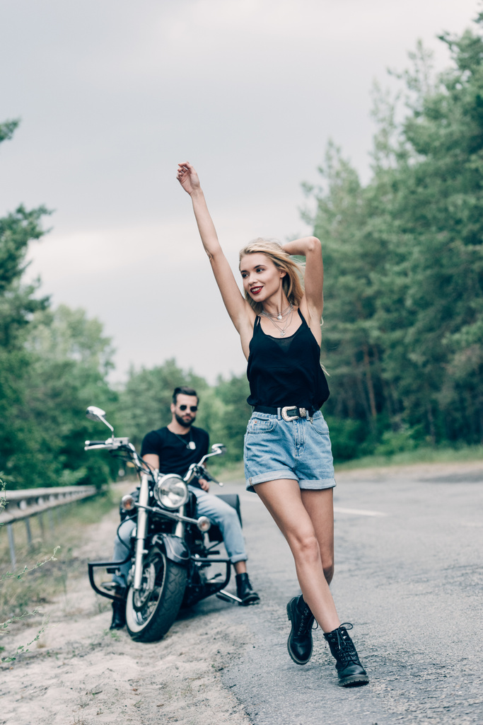 selectieve focus van gelukkige jonge vrouw die op weg loopt met handen in de lucht in de buurt van vriendje op zwarte motorfiets - Foto, afbeelding