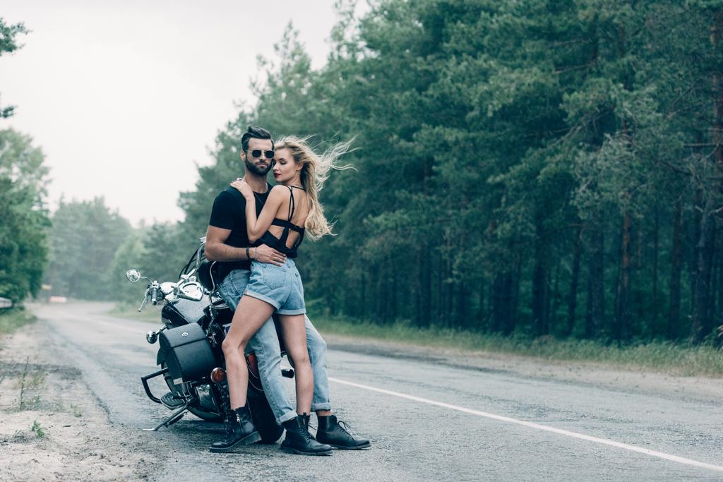 jonge sexy paar motorrijders knuffelen in de buurt van zwarte motorfiets op weg in de buurt van bos - Foto, afbeelding