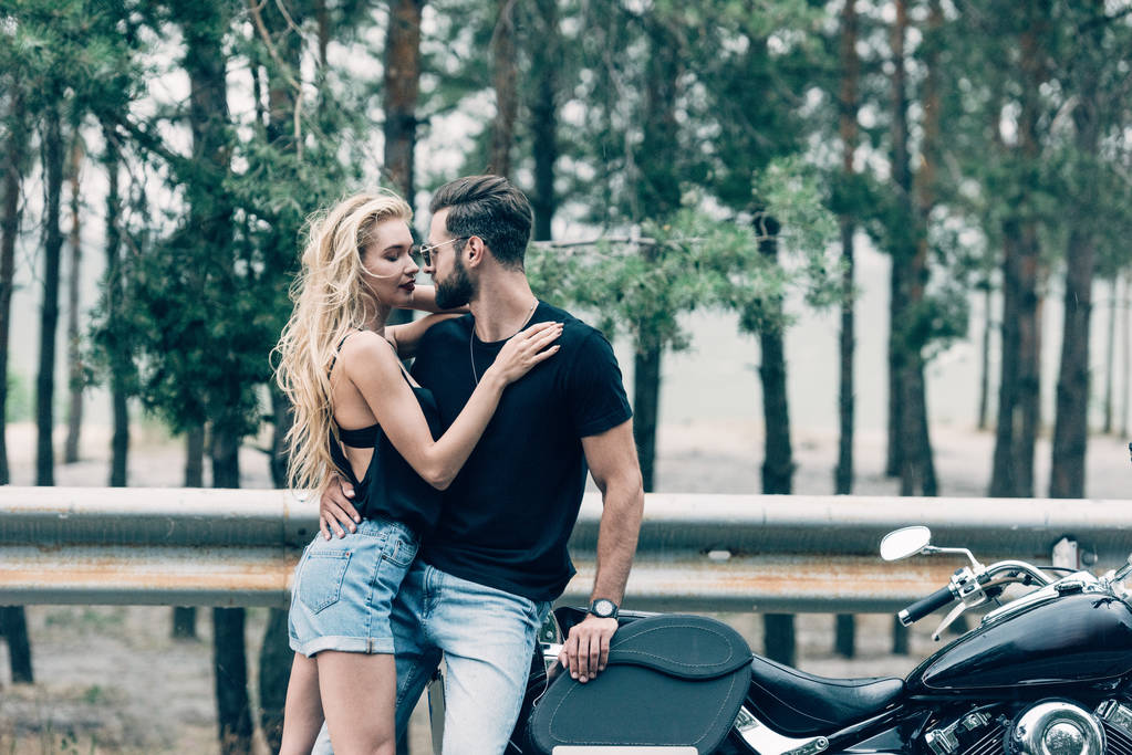 young couple of bikers tenderly embracing near black motorcycle on road near green forest - Photo, Image