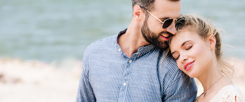 menina loira com os olhos fechados apoiados no ombro namorado, enquanto o homem sorrindo na praia, tiro panorâmico
 - Foto, Imagem