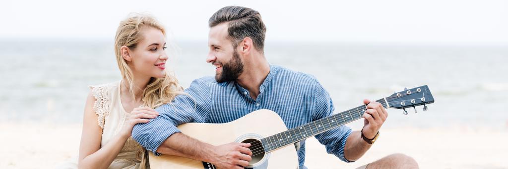 usmívající se krásná mladá žena sedící u šťastného přítele s akustickou kytarou na pláži blízko moře, panoramatický záběr - Fotografie, Obrázek
