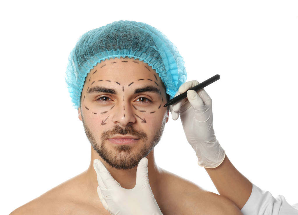Doctor drawing marks on man's face for cosmetic surgery operation against white background - Photo, Image