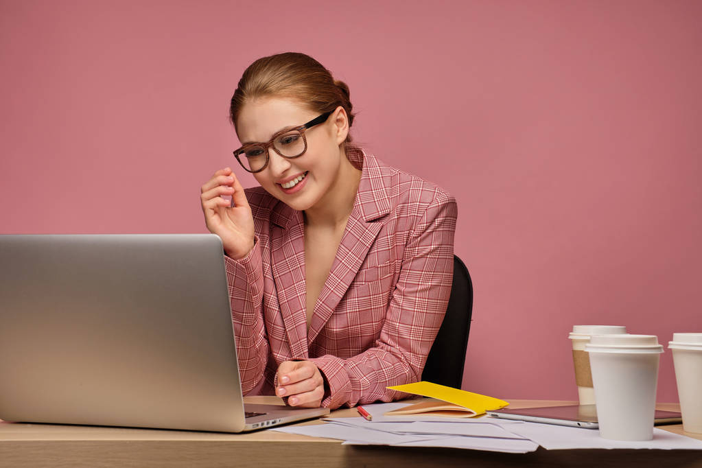 Young woman smiles looking at the laptop sitting at the desk, pink background - Photo, Image