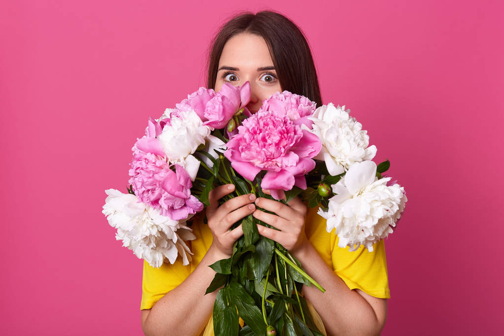 Portret energiczny atrakcyjny Pani z ciemnymi włosami stojący na białym tle na różowym tle, naciskając kwiaty blisko ciała, obejmujące prawie całą część twarzy z piwonie, otwierając oczy szeroko. - Zdjęcie, obraz