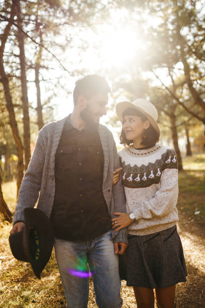 お互いを見て素敵なヒップスターカップル。美しい帽子とセーターを着たカップル。ライフスタイル、公園で晴れた日に2つの遊びの幸せなカップル。若さ、愛、ライフスタイルの概念。秋の夕焼け  - 写真・画像