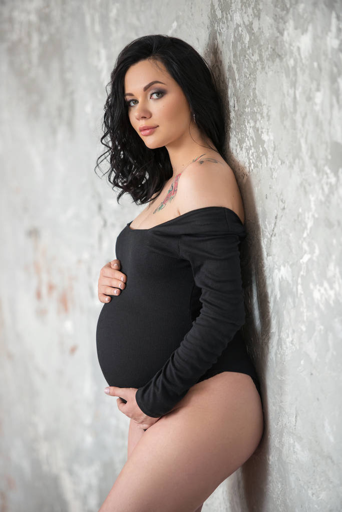 Krásná mladá těhotná brunetka v černém kombinézu se jí pletou na břicho. Stylové těhotenství, ženské zdraví, šťastné mateřství - Fotografie, Obrázek