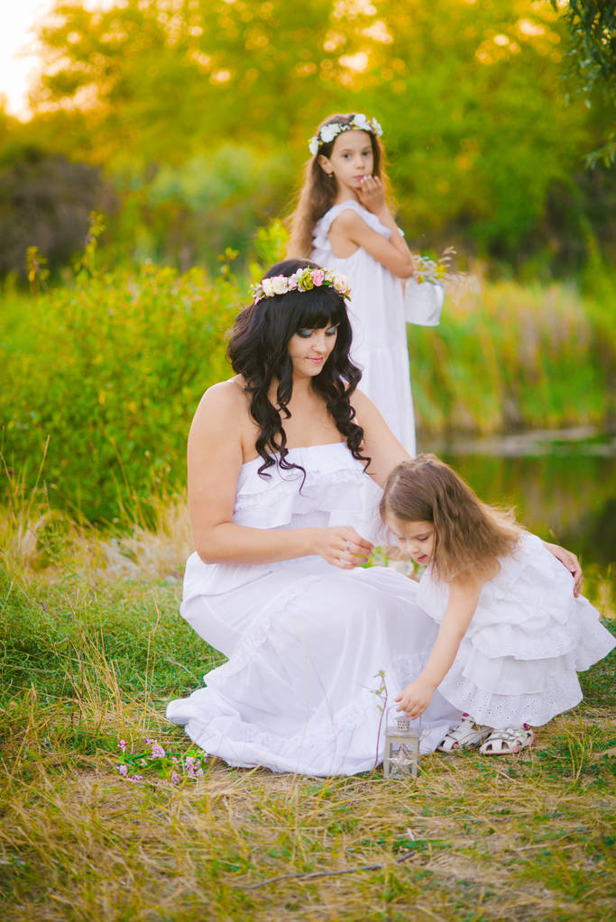 junge Mutter mit ihren Töchtern in weißen Kleidern, die sich in einem Sommerfeld bei Sonnenuntergang am Fluss amüsieren - Foto, Bild