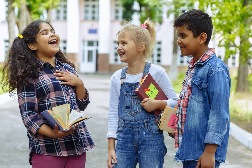 学校に戻れ学校の前で抱き合って笑うバックパックを持つ3人の友人をクローズアップ。校庭で楽しむ学校の子供たちの混合人種グループ. - 写真・画像