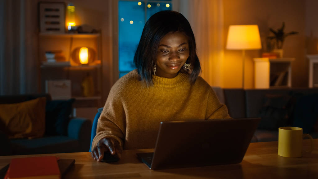 Retrato de una hermosa chica afroamericana sonriente usando un ordenador portátil mientras está sentada en su escritorio en casa. Por la noche, la mujer creativa trabaja en una computadora en su acogedora sala de estar
. - Foto, Imagen