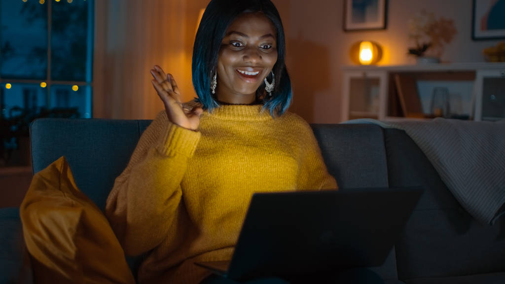 Retrato de una hermosa chica negra sonriente en casa, sentada en un sofá usando un ordenador portátil para hacer una videollamada, dice hola. En la noche chica habla con familiares y amigos utilizando la cámara web de la computadora
. - Foto, Imagen