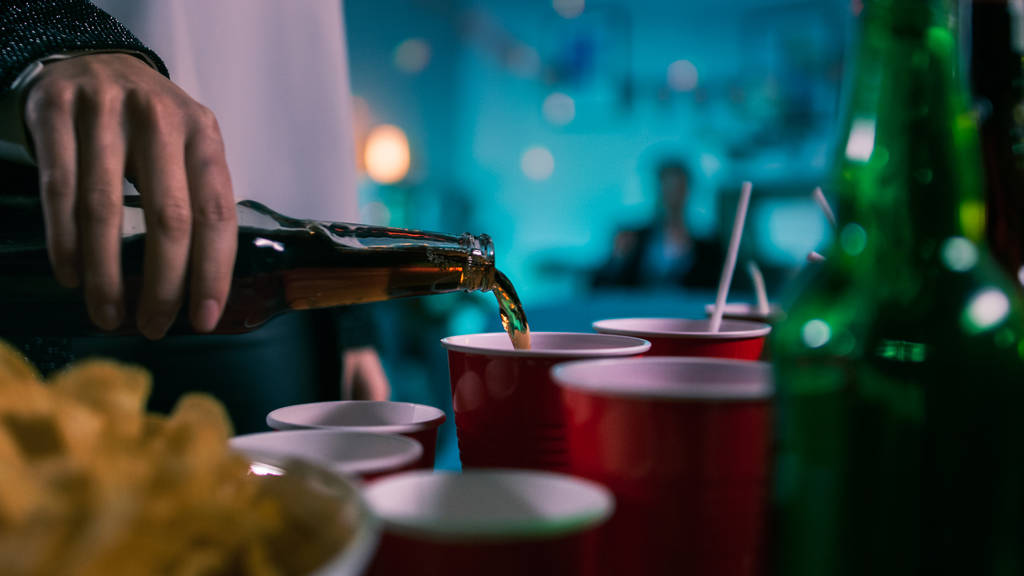 На дикій вечірці: людина наливає напій з пляшки в червону чашку. У фоновому режимі розмиті люди танцюють. - Фото, зображення