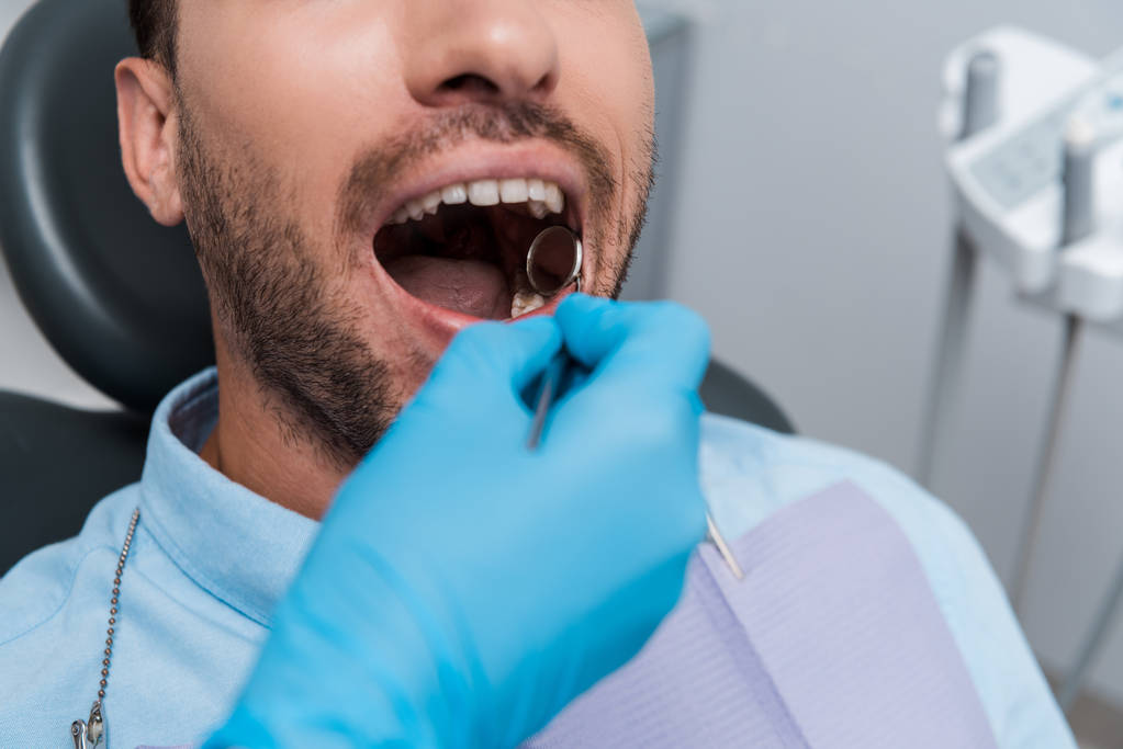 visão recortada do dentista segurando espelho dentário na boca do paciente barbudo
  - Foto, Imagem