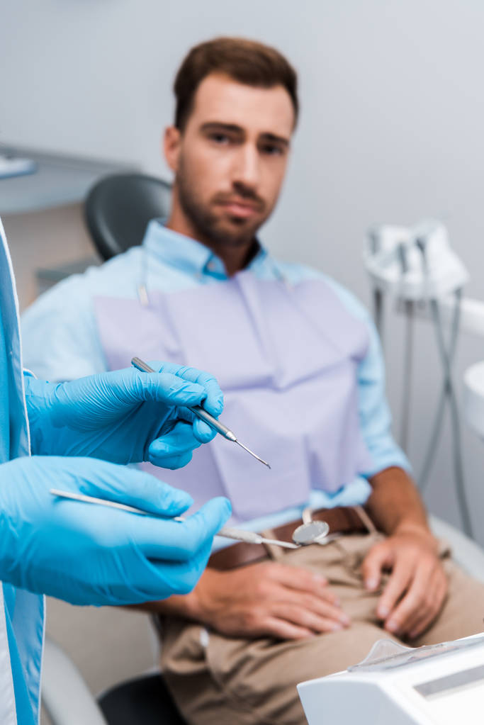 επιλεκτική εστίαση του οδοντιάτρου σε γάντια από μπλε λατέξ που κατέχουν οδοντιατρικά όργανα κοντά στον άνθρωπο  - Φωτογραφία, εικόνα