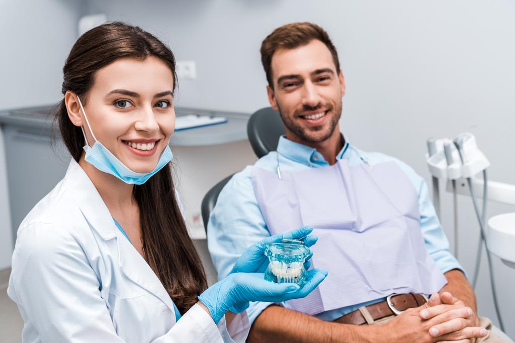 επιλεκτική εστίαση του ελκυστικού οδοντίατρου σε γάντια λατέξ κρατώντας μοντέλο δοντιών κοντά σε ασθενή με σφικτά χέρια  - Φωτογραφία, εικόνα
