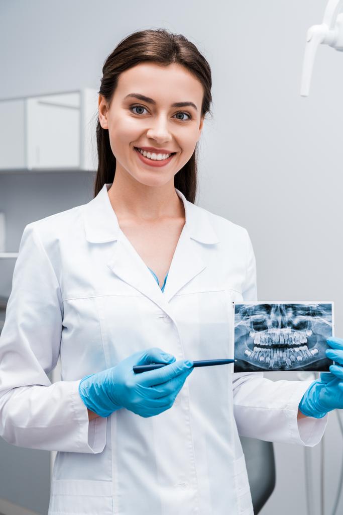 陽気な歯科医がX線の近くでペンを持ち、診療所で微笑む  - 写真・画像