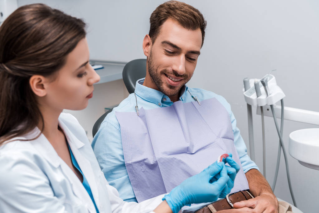 επιλεκτική εστίαση του χαρούμενο ασθενή καθισμένος κοντά στον οδοντίατρο και κοιτάζοντας το μοντέλο των δοντιών στην κλινική  - Φωτογραφία, εικόνα
