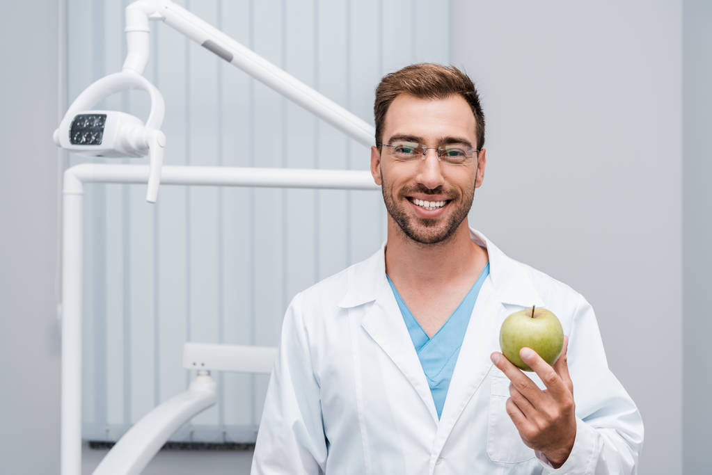 happy bearded doctor in glasses holding green and tasty apple  - Photo, Image