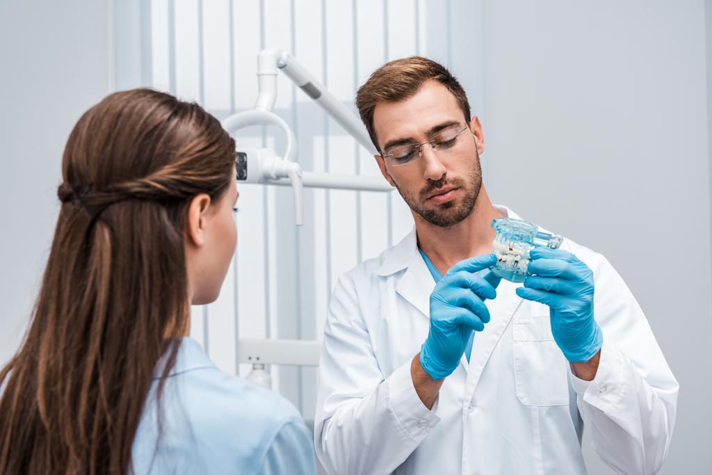 επιλεκτική εστίαση του μουσάτο οδοντίατρο σε ποτήρια κρατώντας μοντέλο δοντιού κοντά στον ασθενή - Φωτογραφία, εικόνα