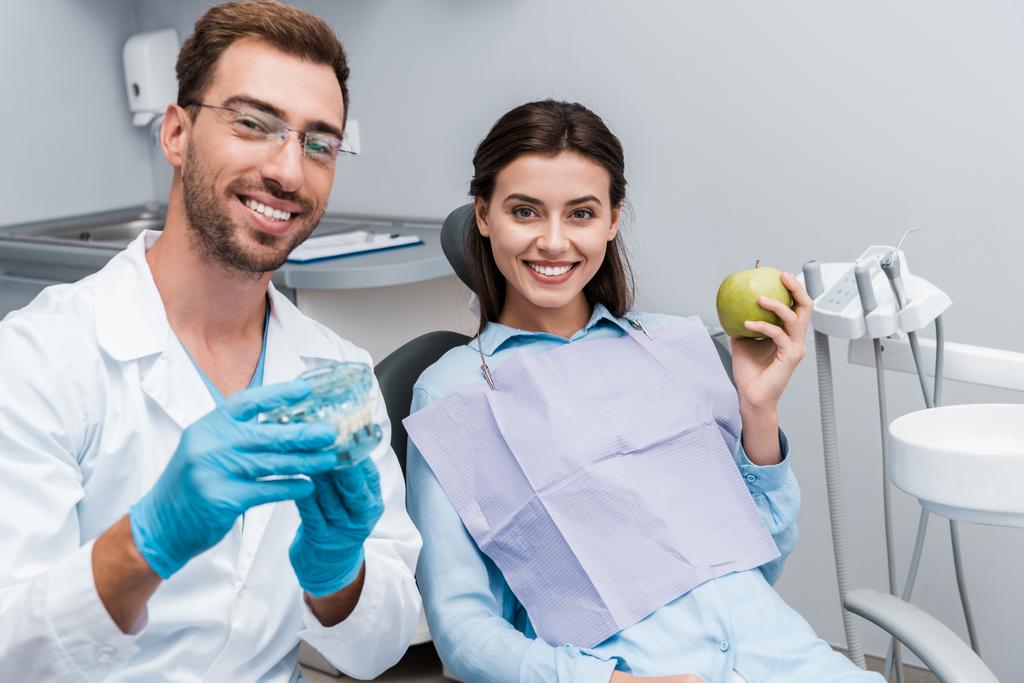селективное внимание веселого стоматолога в очках, держащего в руках модель зубов рядом с счастливой женщиной с зеленым яблоком
  - Фото, изображение
