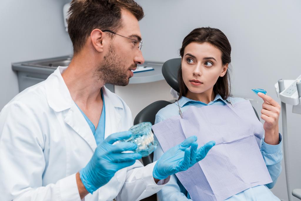 リテーナーを持つ女性の近くでジェスチャー眼鏡でハンサムな歯科医の選択的な焦点  - 写真・画像