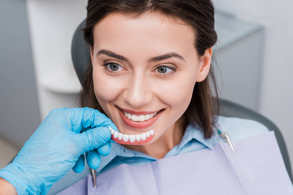 περικομμένη όψη του οδοντιάτρου κρατώντας πρόθεση κοντά ευτυχισμένο κορίτσι στην οδοντιατρική κλινική  - Φωτογραφία, εικόνα