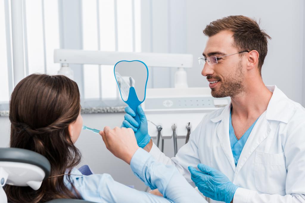 selectieve focus van vrolijke tandarts Holding spiegel in de buurt van patiënt poetsen tanden  - Foto, afbeelding