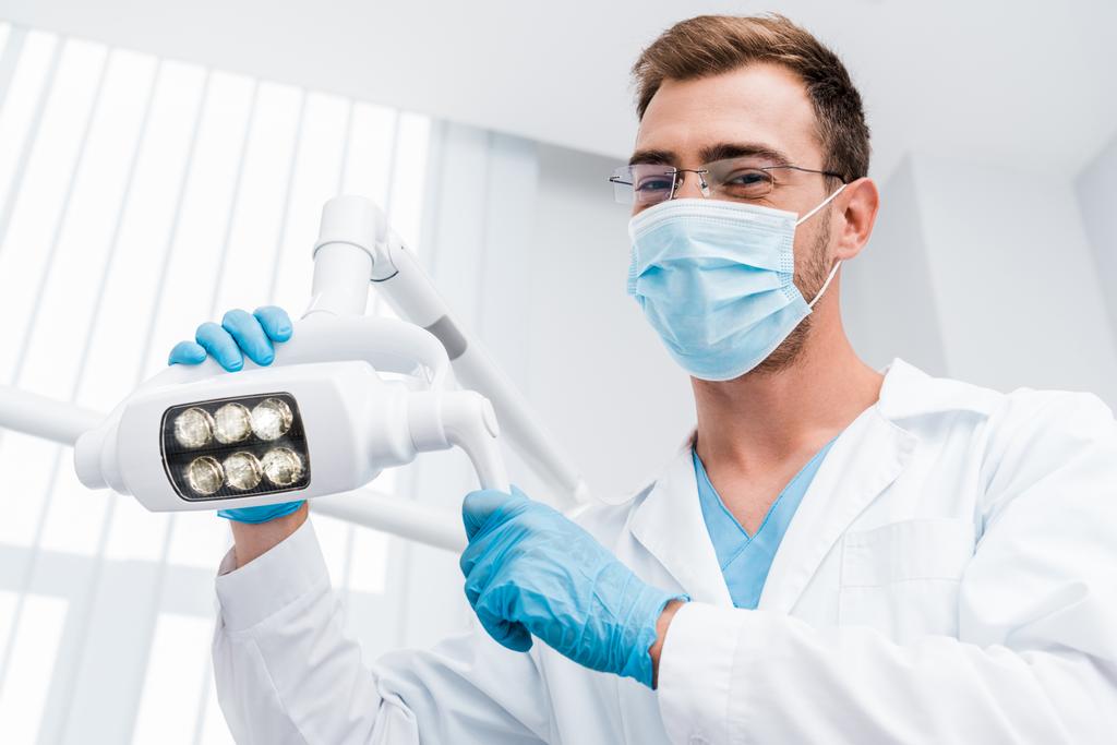 niski kąt widzenia dentysta w okularach i maska medyczna dotykając lampy medycznej i patrząc na kamerę  - Zdjęcie, obraz