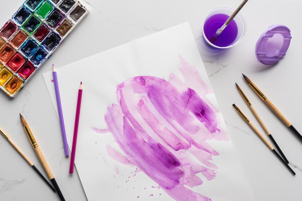 top view of purple watercolor brushstrokes on white paper near paintbrushes, paints and colored pencils on marble white surface - Photo, Image